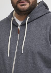 Felpa grigia con zip realizzata in tessuto morbido, dotata di coulisse con dettagli crema e zip in metallo. Dettaglio in pelle ben visibile.