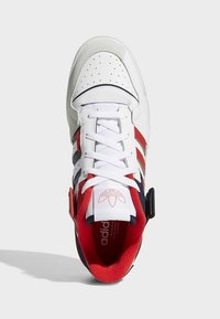 Baskets blanches Adidas avec des rayures rouges et noires, des lacets blancs et une doublure intérieure rouge, vues de dessus sur un fond uni.