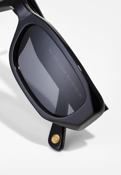 Ochelari de soare din acetat negru cu lentile întunecate. Prezintă un design unghiular și un detaliu aurit în formă de raze de soare pe bradă. Branding gravat pe lentilă.