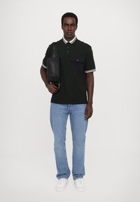 Mørk grønn polo-skjorte med hvitstripet krage, enkelt brystlomme. Lys blå denimjeans og hvite joggesko. Svart crossover-veske.