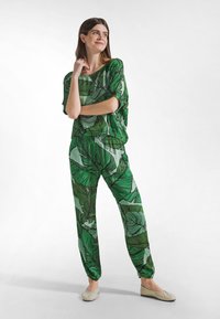 Zielony kombinezon w liściasty wzór, wykonany z lekkiej tkaniny, o luźnym kroju, szerokich rękawach i elastycznych mankietach na spodniach.