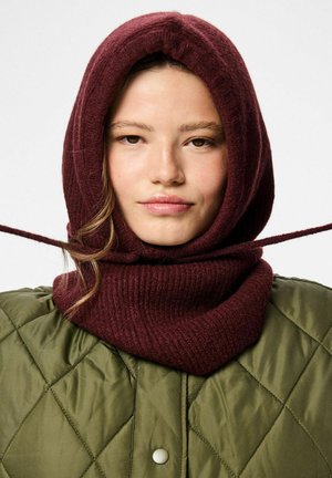 Burgunderfarbene gestrickte Kapuze mit geripptem Muster, kombiniert mit einer grünen gesteppte Jacke mit Rautenmuster und Druckknöpfen.