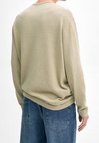 Maglione color khaki chiaro con maniche lunghe e scollatura tonda, abbinato a jeans di denim blu con tasche posteriori e una vestibilità comoda.