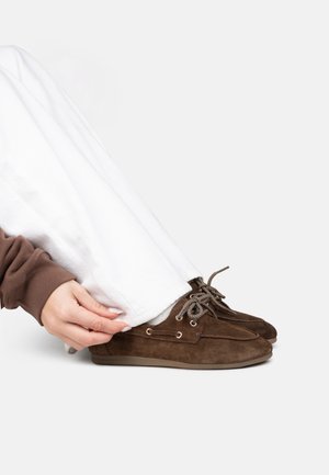 Personne portant un pantalon blanc et des mocassins en daim marron, ajustant la jambe du pantalon en étant assise devant un fond uni.