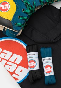 Večbarvni skate čevlji z rumenimi, modrimi in zelenimi detajli ter dvema paroma vezalk (črnimi in teal), prikazani na črni površini.