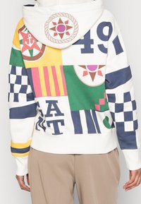 Osoba w wielokolorowej bluzie z kapturem, ozdobionej wzorami marynistycznymi i geometrycznymi, cyframi oraz literami, w połączeniu z beżowymi spodniami, widok z tyłu.