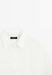 Camicia bianca in lino con colletto classico, chiusura con bottoni sul davanti e texture leggera. Presenta l'etichetta del marchio sul lato interno del colletto.