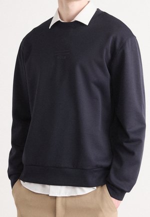 Homme portant un sweat-shirt bleu marine par-dessus une chemise blanche à col, avec un pantalon kaki, les mains dans les poches, sur un fond uni.