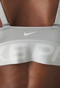 Svetlo siva športna modrček z širokim pasom, ki prikazuje bel logotip "NIKE". Gladka tekstura, nastavljive naramnice in minimalistična zasnova.