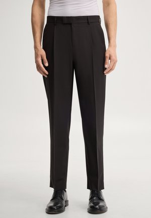 Man draagt zwarte, op maat gemaakte nette broek met vouwen aan de voorkant en zwarte leren veterschoenen op een egale lichte achtergrond.