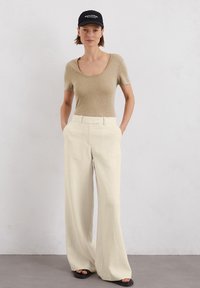 Beige getailleerde top, brede crèmekleurige broek, zwarte pet en donkere sandalen. Zachte textuur en ontspannen pasvorm met een soepele, natuurlijke kleurenpalet.
