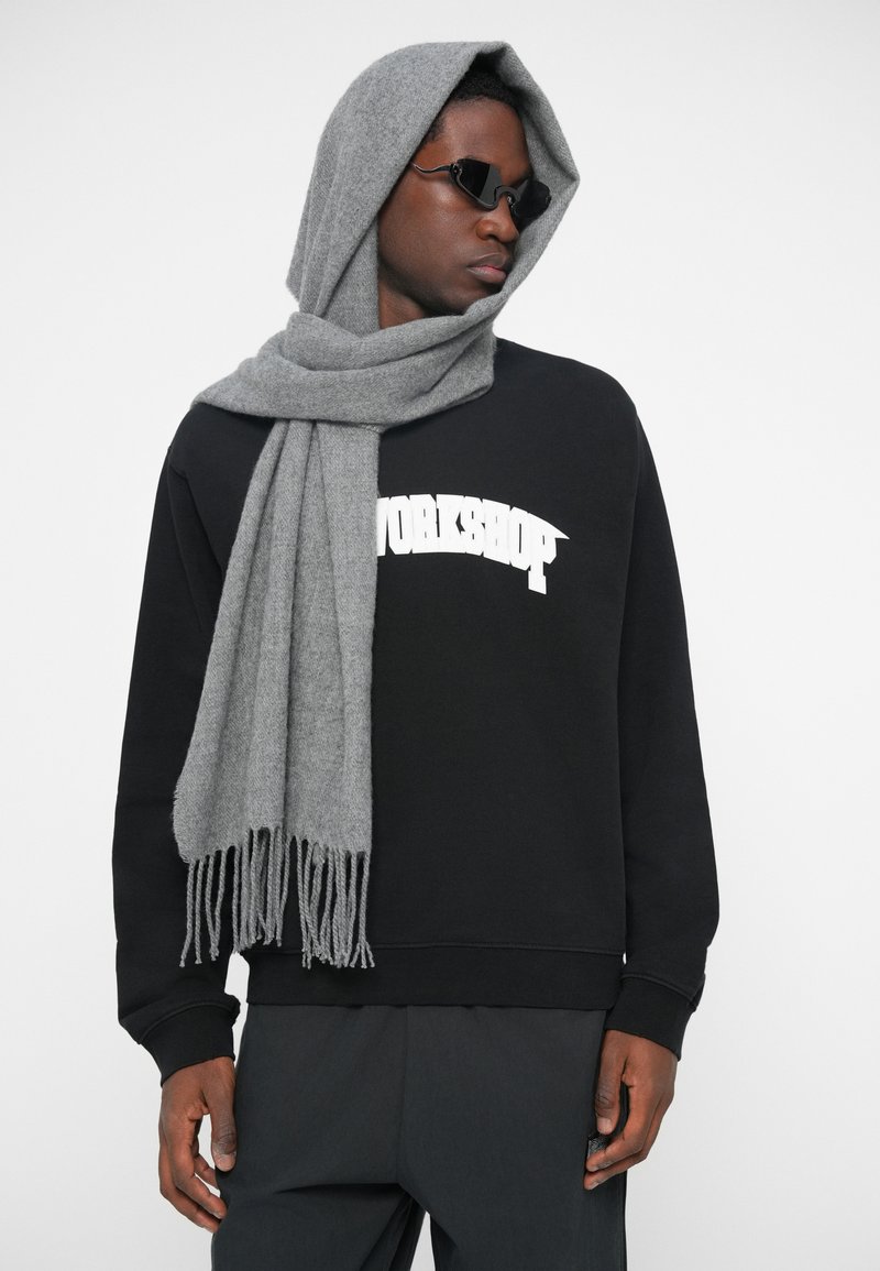 Man in een zwarte sweatshirt met de tekst "WORKSHOP", zwarte broeken, donkere zonnebril en een grijze sjaal over zijn hoofd en schouder gedrapeerd.