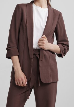Femme portant un blazer marron et un pantalon taille haute assorti avec ceinture, associé à un t-shirt blanc, sur un fond gris uni.