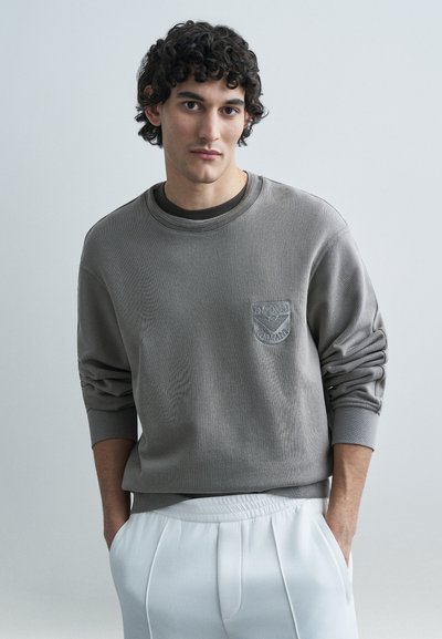 Grå sweatshirt med ribbet tekstur, rund hals og broderet logo på venstre bryst. Kombineret med hvide plisserede bukser.