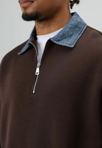 Bruine sweatvest met rits en een denim kraag. Voorzien van een zilveren rits en een zachte textuur, met een minimalistisch ontwerp.