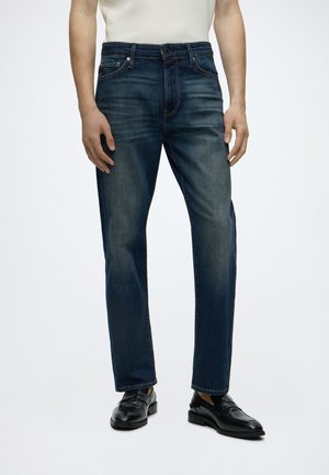 Homme portant un jean bleu foncé et des mocassins en cuir noir, debout les mains détendues le long du corps, devant un fond uni.