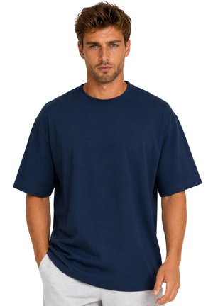 Man met kort bruin haar, draagt een loszittend marineblauw T-shirt en lichtgrijze broek, staat met handen in de zakken tegen een witte achtergrond.