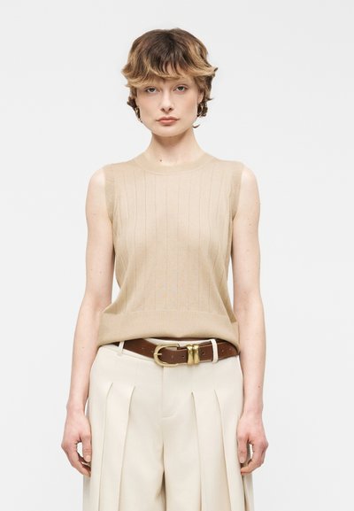 Femme aux cheveux courts portant un haut beige sans manches côtelé, un pantalon plissé crème et une ceinture marron avec une boucle dorée, debout devant un fond blanc.