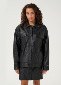 Melnā ādas jaka ar strukturētu dizainu, papildināta ar metāla kniedēm ap apkakli un krūškurvja kabatām, kā arī ar pogu aizdari.