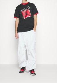 Černé tričko s červenými akcenty a grafikou "Flight" na přední straně, spárované s volnými bílými cargo kalhotami a černými/červenými teniskami.