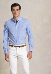 Gai zila krekla ar pogām, kas izgatavots no kokvilnas, ar piegriezumu, apvienots ar baltām džinsām un brūnu ādas jostu. Uz krūtīm ir smalkais logs.