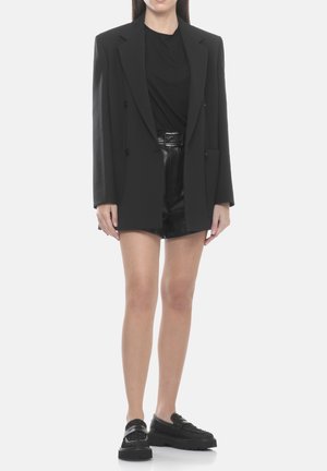 Donna che indossa un blazer nero oversize, una t-shirt nera, pantaloncini in pelle neri a vita alta e mocassini neri chunky, in piedi su sfondo bianco.