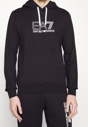 Ensemble sweat à capuche et pantalon noir avec logo blanc Emporio Armani EA7 sur la poitrine et la cuisse gauche, porté par une personne debout devant un fond blanc.