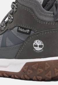 Šedá vodotěsná turistická bota s texturovaným svrškem, kovovými detaily, logem Timberland a robustní gumovou podrážkou s trojúhelníkovými vzory.