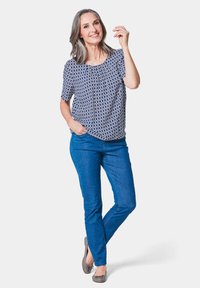 Blauwe blouse met patroon en korte mouwen, gedragen met blauwe jeans en grijze ballerina's. Model staat casual met één hand in de zak.