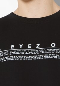 Černé tričko s kulatým výstřihem, na kterém jsou bílé potisknuté názvy písniček a jejich délky uspořádány do kruhu na oblasti hrudníku.