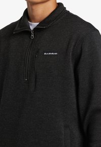 Tmavo šedý fleece pulóver s vysokým golierom, polovičným zipsom na prednej strane a vreckom na hrudi. Obsahuje logo na ľavej strane. Textúrovaná tkanina.