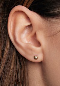 Złoty kolczyk w kształcie serca, o błyszczącej powierzchni, małego rozmiaru, noszony na płatku ucha, eksponujący fakturę i krzywiznę wzoru.