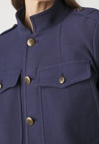 Chaqueta azul marino fabricada en tejido texturizado, con botones dorados y dos bolsillos en el pecho, con un cuello alzado y un ajuste entallado.