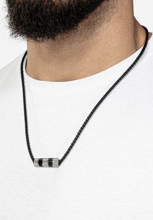Black braided necklace with a metallic cylindrical pendant featuring engraved patterns and black accents, set against a white t-shirt background.