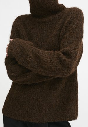 Person wearing a dark brown fuzzy turtleneck sweater with long sleeves, arms crossed over the chest against a plain background.