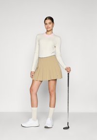 Pull en crème à côtes avec col rose, jupe plissée beige, chaussures blanches et chaussettes crème, tenant un club de golf. Design simple et couleurs neutres.
