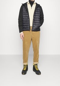 Svart pufferväst över en ljusbeige tröja, parat med beigefärgade kordbyxor och svarta stövlar med gula snören. Enkel, funktionell design.