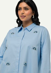 Camicia azzurra chiara in cotone, con intricate decorazioni ricamate blu e argento, colletto a bottoni e maniche lunghe.