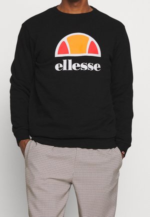 Man wearing black ellesse sweatshirt with orange and red semi-circle logo, paired with black and white checkered pants, hands in pockets.