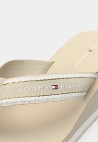 Béžové žabky s textilními pásky zdobenými zlatými detaily. Na pásku je potisk s logem "TOMMY HILFIGER". Texturovaná stélka a hladká podešev.
