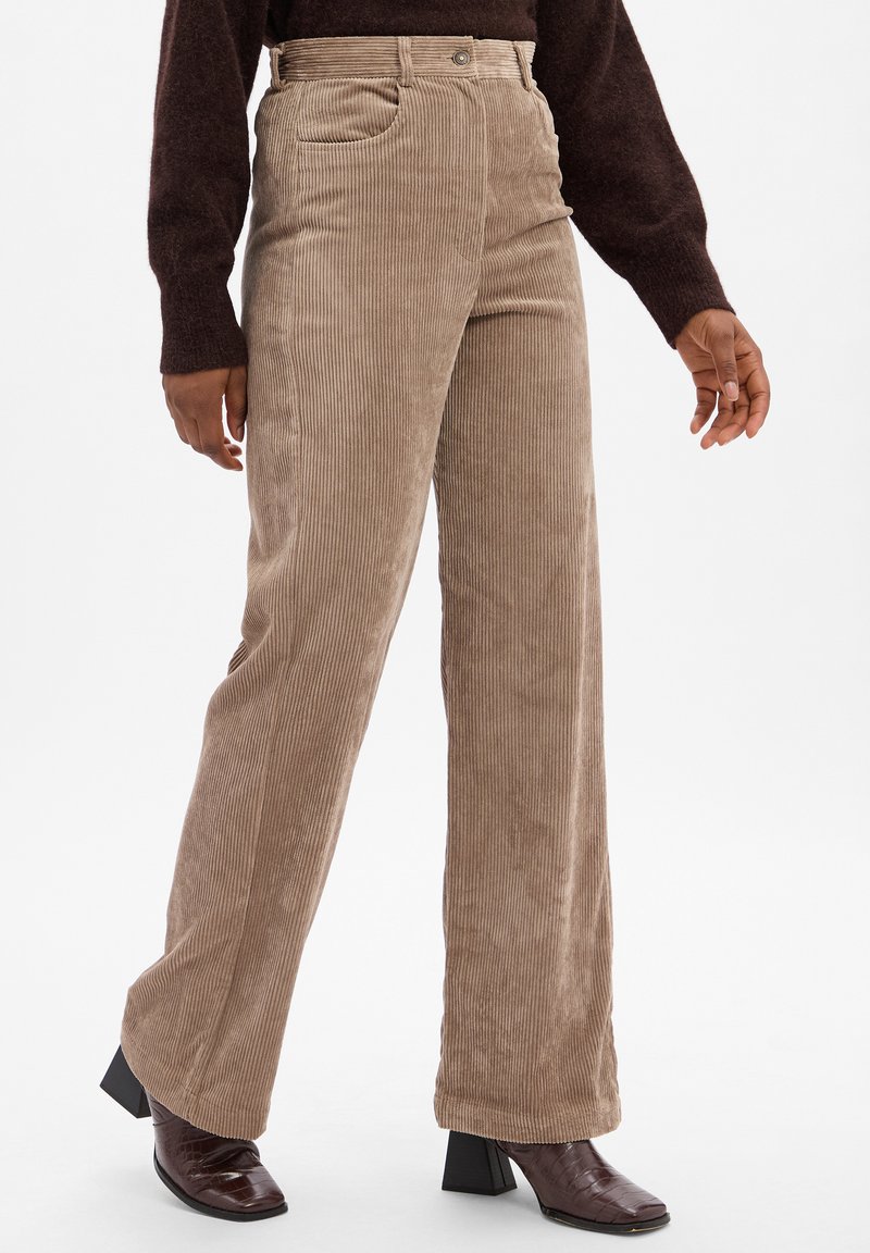 Pantalons large en velours côtelé marron clair avec des côtes verticales. Il présente des poches avant, une fermeture à bouton et une texture lisse.