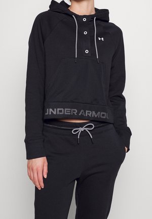 Person trägt schwarzen Under Armour Hoodie mit vorderer Netz-Tasche und schwarze Sweatpants mit Kordelzug vor einfarbigem Hintergrund.