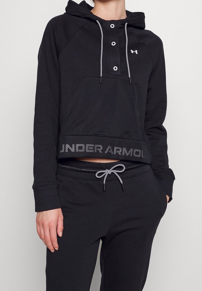 Personne portant un sweat à capuche noir Under Armour avec poche en mesh à l’avant et un pantalon de jogging noir avec cordon, sur fond uni.