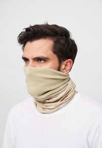 Beige fleece halsgjerde med et glatt, stretchy innerlag og et teksturert ytre lag, båret rundt halsen og delvis dekkende ansiktet.