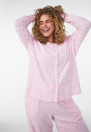Mujer sonriente con cabello rizado que lleva un pijama rosa con pequeños corazones rojos, posando con las manos detrás de la cabeza sobre un fondo blanco.
