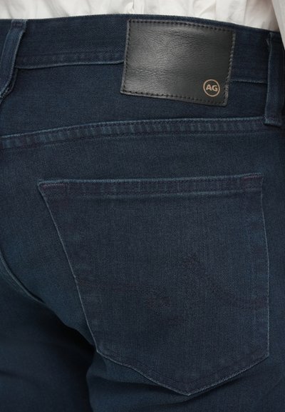 Σκούρα μπλε τζιν παντελόνια με λεία υφή, πίσω μπεζ τσέπη και μια μαύρη δερμάτινη ετικέτα πίσω, εντυπωμένη με το "AG."