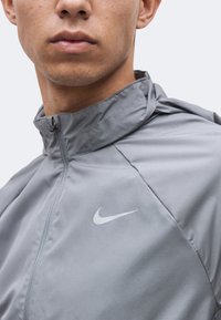 Νέος άνδρας φορά ένα ανοιχτό γκρι μπουφάν Nike με φερμουάρ, ψηλό γιακά και ορατό λευκό λογότυπο Nike Swoosh στο στήθος.