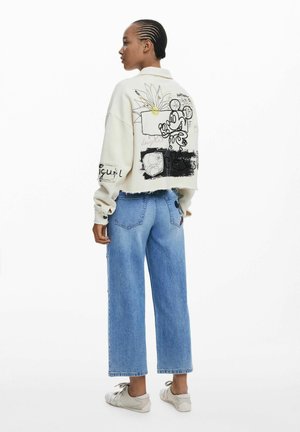 Afgekorte beige sweatshirt met grafische prints in zwart en groen; hoge lichtblauwe wijde jeans; grijze sneakers.