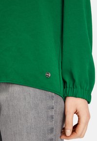 Green pullover with long sleeves featuring gathered cuffs and a metallic logo accent on the hem, paired with gray denim.