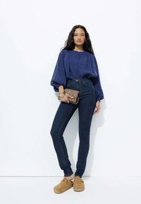 Blouse marine à manches bouffantes, jeans en denim foncé taille haute, et sabots en suède beige. Tenant une pochette en cuir imprimé léopard marron.